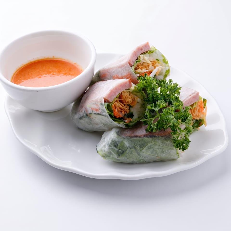 Three fresh spring rolls with ham, vegetables, and herbs, served with a side of orange dipping sauce.